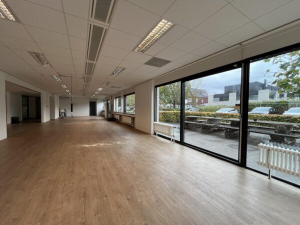 Leegstaand kantoorruimte aan de Burgemeester Verderlaan met grote ramen en uitzicht op een parkeerplaats en straat.